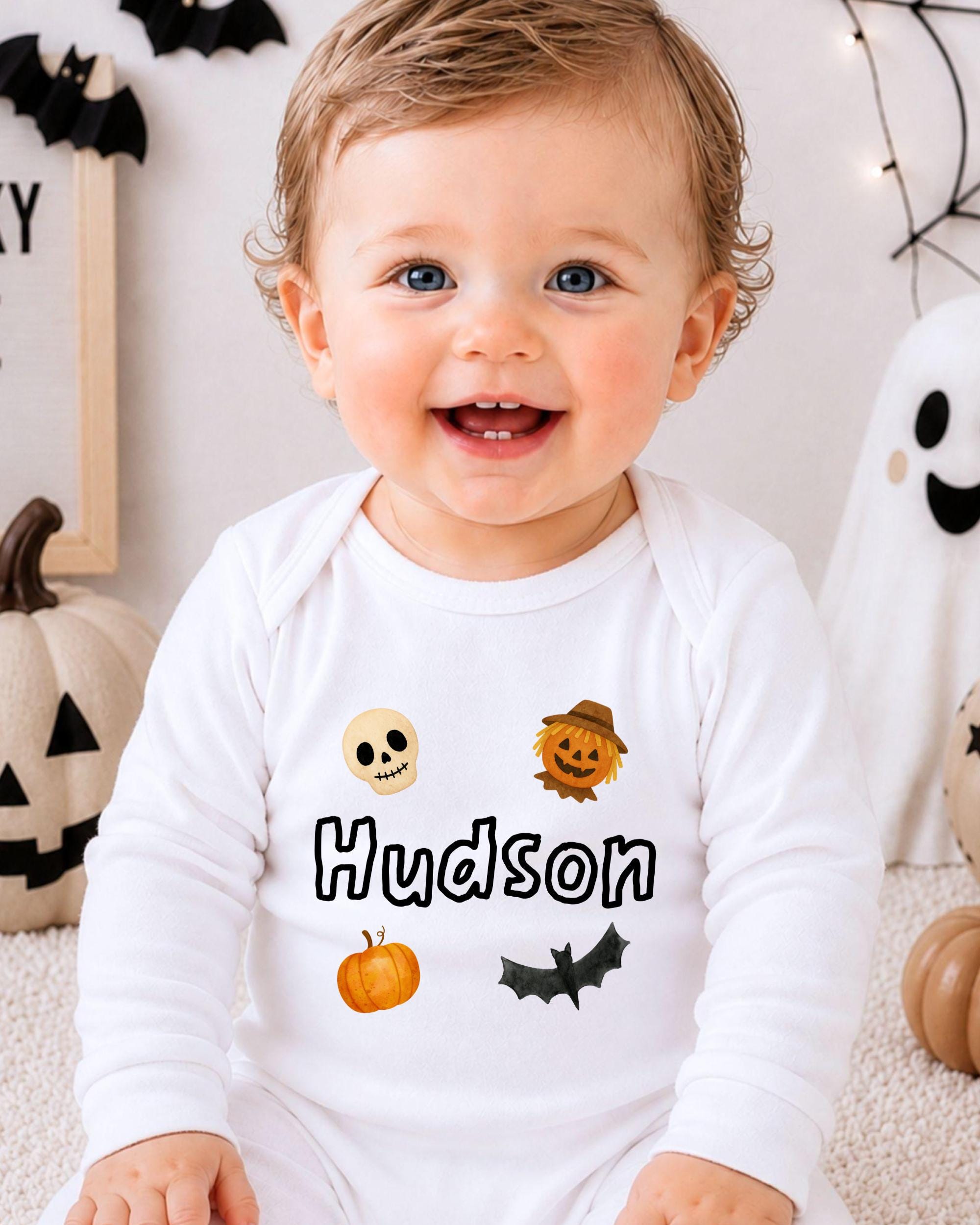 A smiling baby boy wearing a white onesie with Halloween-themed designs and the name "Hudson" printed on it.