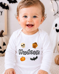 A smiling baby boy wearing a white onesie with Halloween-themed designs and the name "Hudson" printed on it.