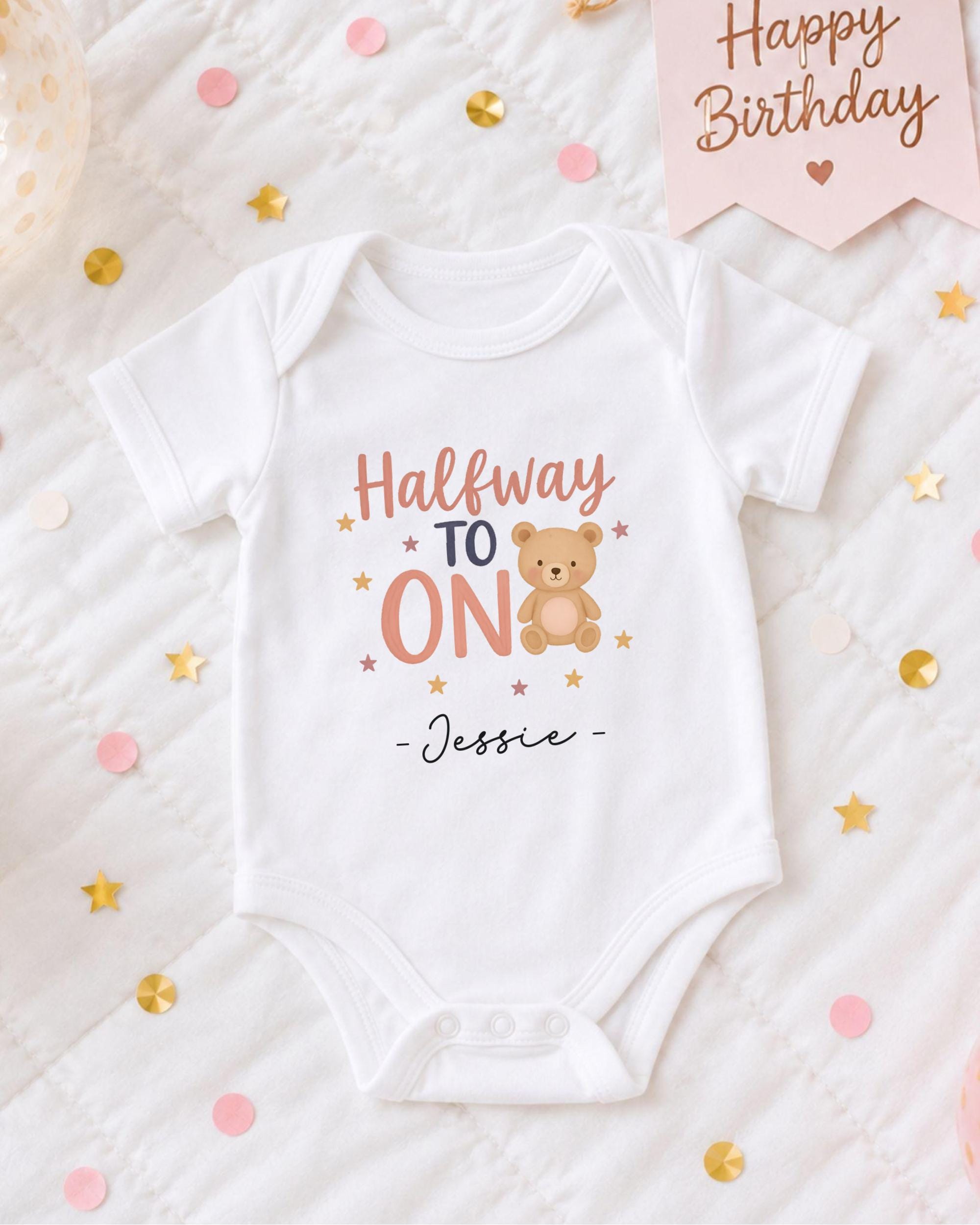 A white onesie with a teddy bear and the text "Halfway to ON" printed on it, along with the name " Jessie" written below. The onesie is placed on a bed with confetti and a birthday card.