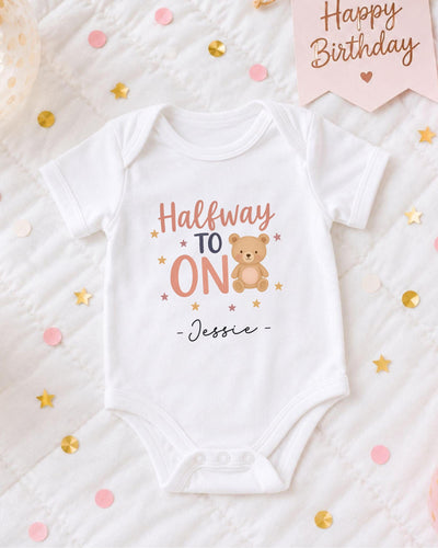 A white onesie with a teddy bear and the text "Halfway to ON" printed on it, along with the name " Jessie" written below. The onesie is placed on a bed with confetti and a birthday card.