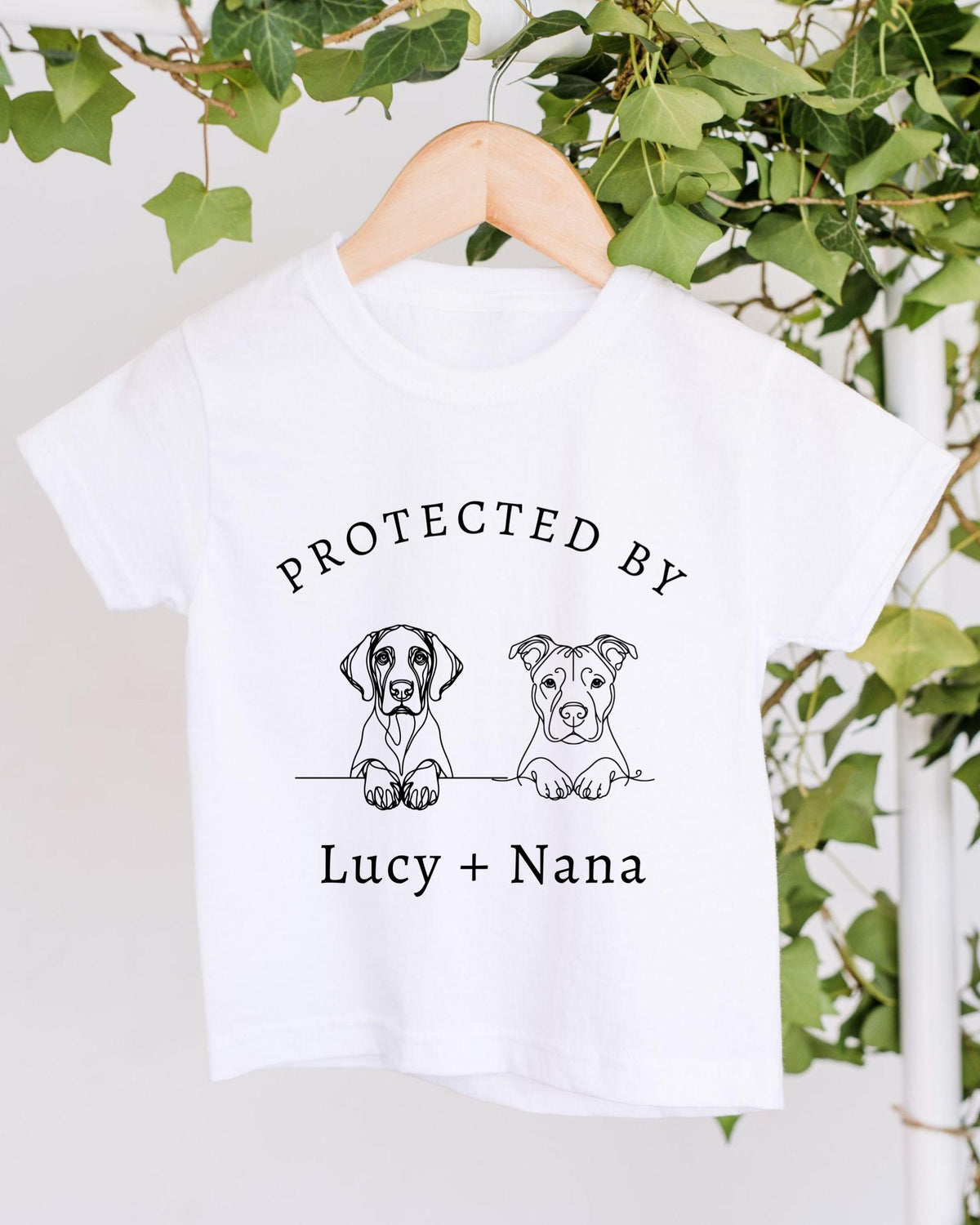 A white t-shirt with a graphic design featuring two cartoon dogs, one labeled "Lucy" and the other "Nana", along with the text "PROTECTED BY Lucy + Nana".