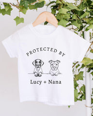 A white t-shirt with a graphic design featuring two cartoon dogs, one labeled "Lucy" and the other "Nana", along with the text "PROTECTED BY Lucy + Nana".