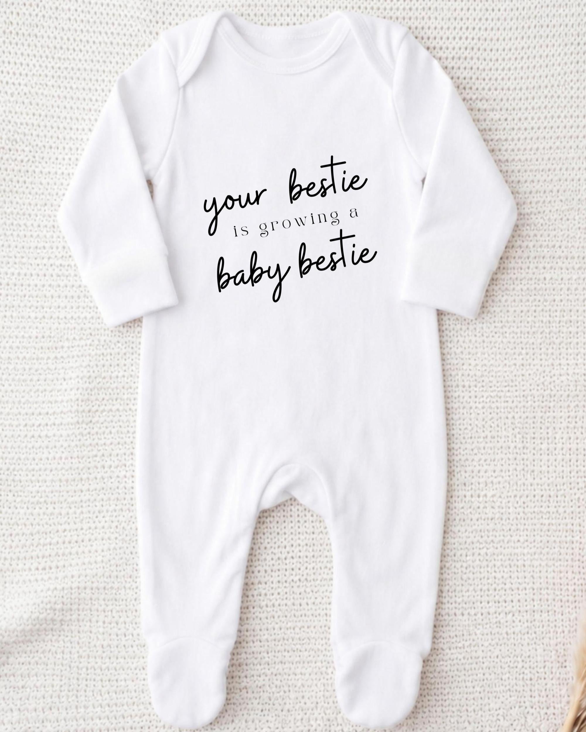 A white onesie with the text "your bestie is growing a baby bestie" printed on it.