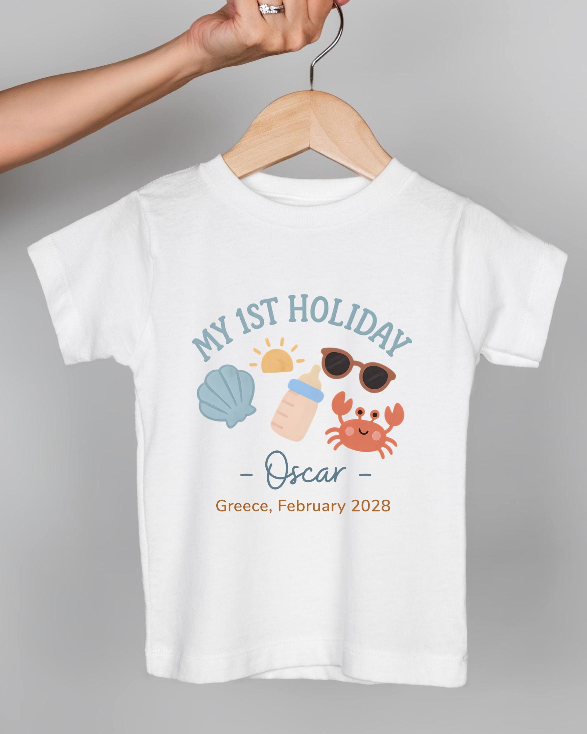 A white t-shirt with a graphic design featuring a baby bottle, sunglasses, a shell, and a crab, along with the text "My 1st Holiday Oscar - Greece, February 2028".