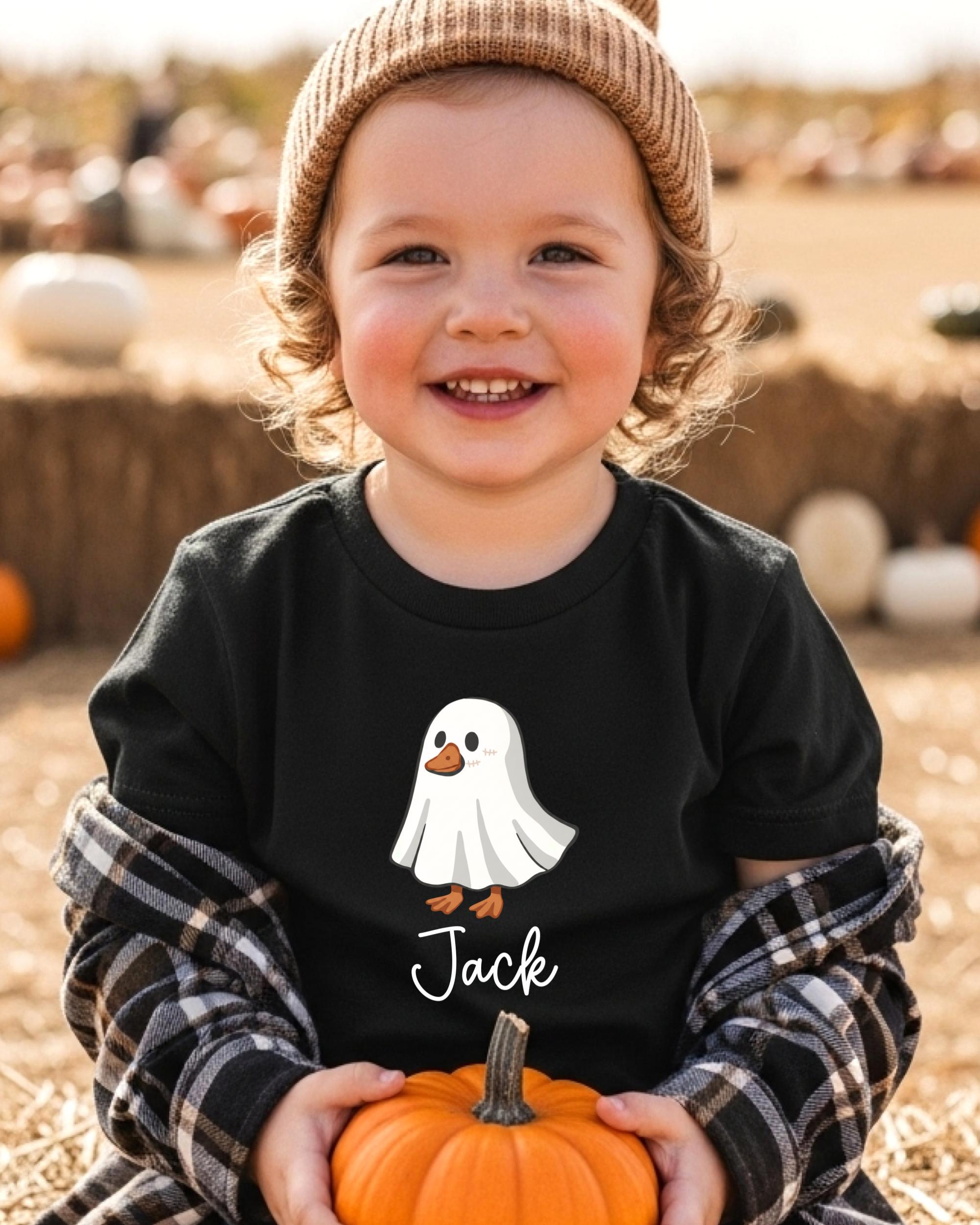 A young child wearing a black t-shirt with a ghost and the name "Jack" printed on it, holding a small orange pumpkin.