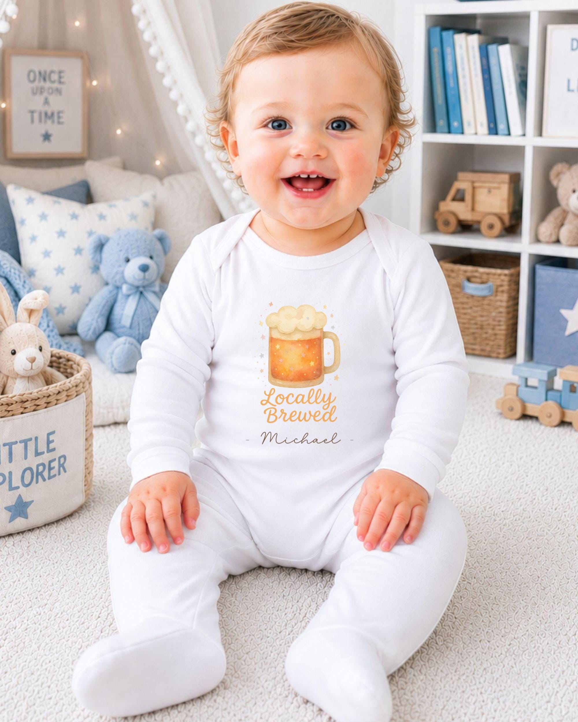 A smiling baby boy wearing a white onesie with a beer graphic and the name "Michael" printed on it, sitting on a carpeted floor in a room with bookshelves and stuffed animals.
