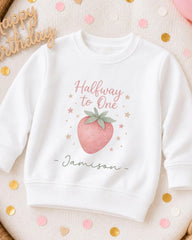 A white sweatshirt with a pink strawberry graphic and the text "Halfway to One" printed on it, along with the name "Jasison" written below. The sweatshirt is surrounded by confetti.