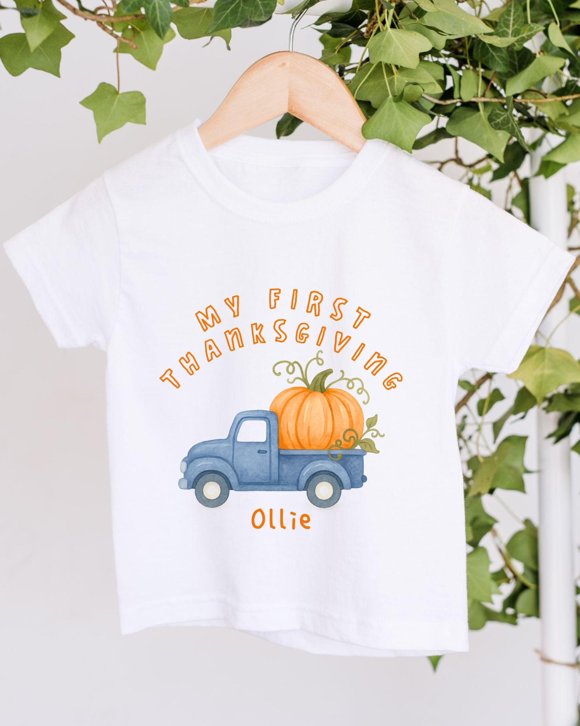 A white t-shirt with a graphic design featuring a blue truck carrying a large pumpkin, along with the text "My first Thanksgiving Ollie" printed on it.