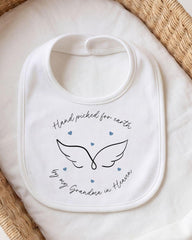 A white bib with a hand-drawn wing design and the text "Hand picked for earth by my Grandma in Heaven" printed on it.