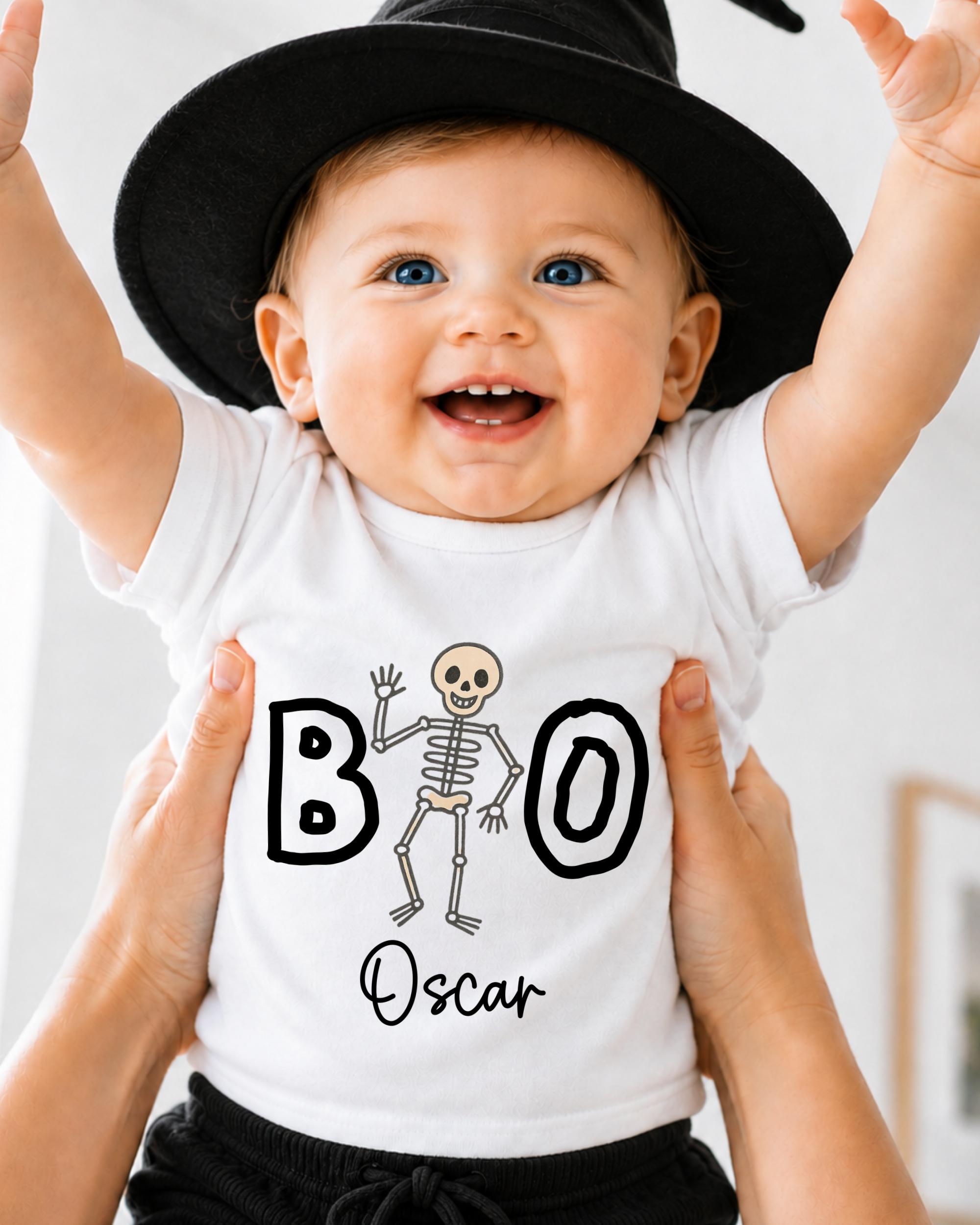 A smiling baby wearing a white t-shirt with a skeleton design and the name "Oscar" printed on it, being held up by someone's hands.