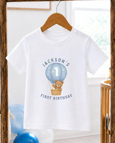 A white t-shirt with a graphic design of a teddy bear in a hot air balloon, along with the text "JACKSON'S FIRST BIRTHDAY".