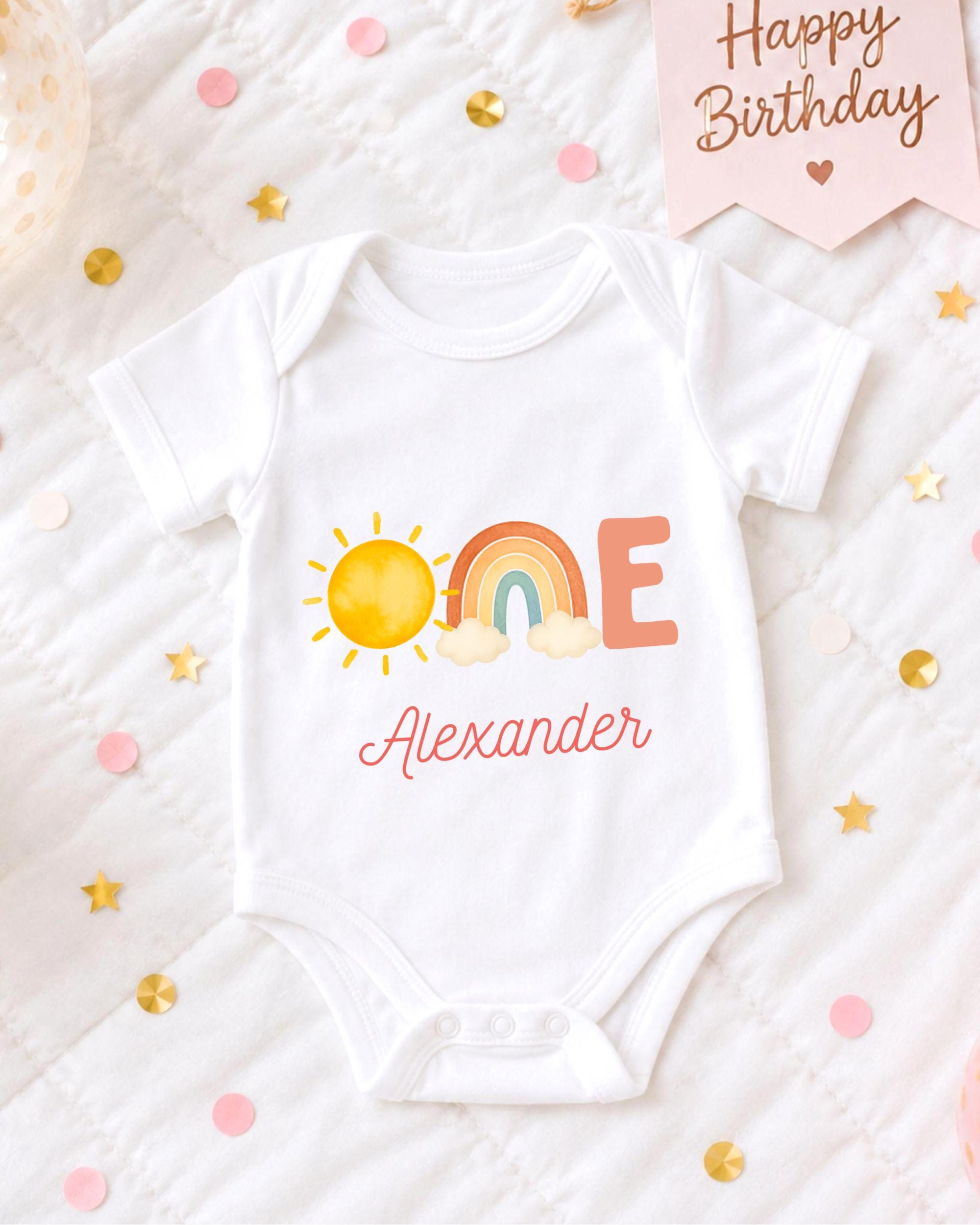 A white onesie with a sun and rainbow design, along with the name "Alexander" printed on it, is displayed on a bed surrounded by confetti.