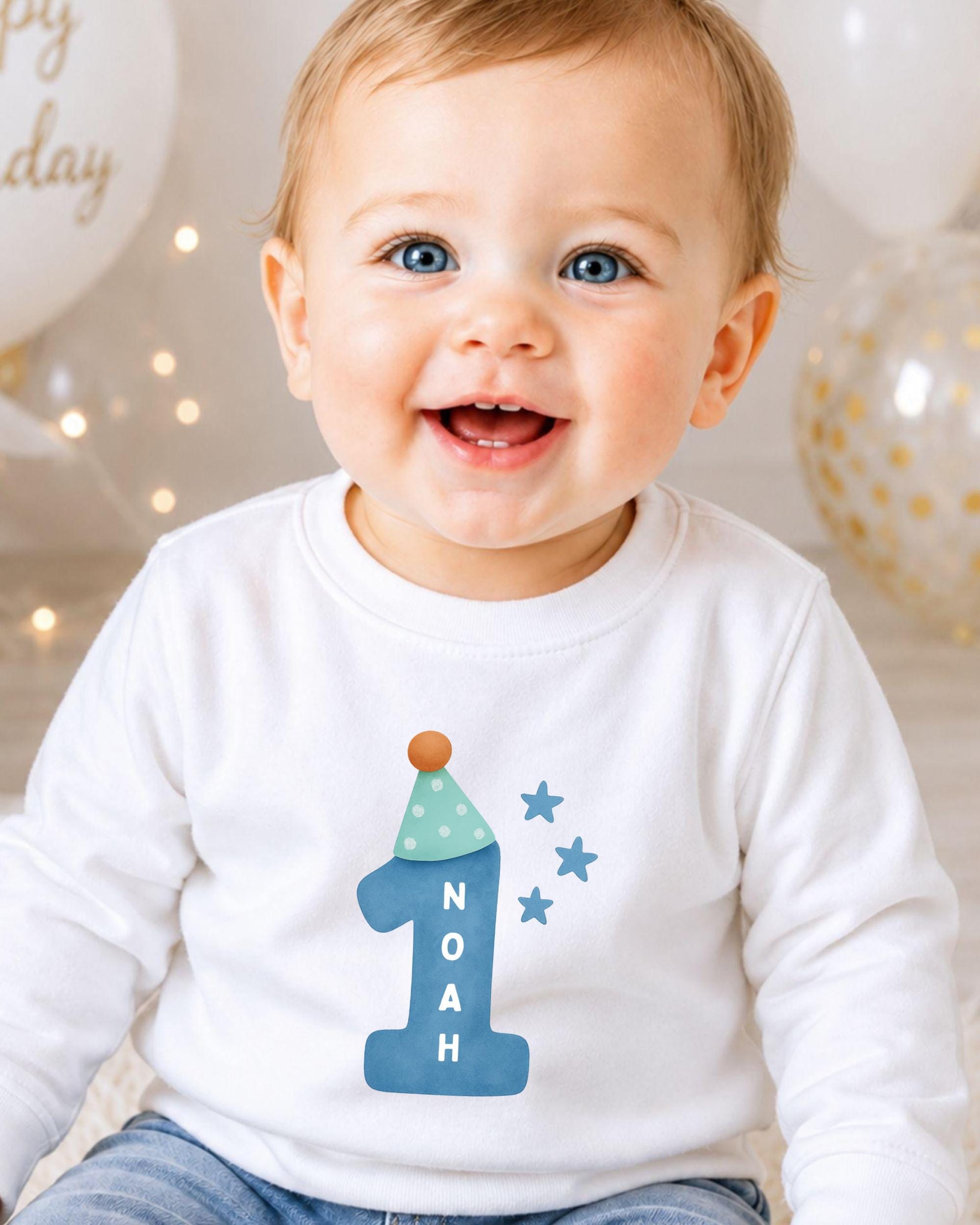 A smiling baby boy wearing a white shirt with a blue number one and stars on it, sitting in front of birthday balloons.
