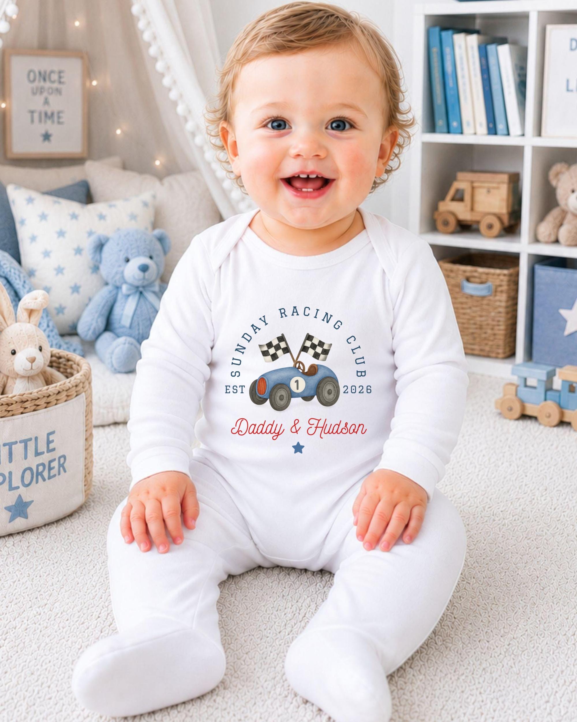 A smiling baby boy wearing a white onesie with a racing club logo sits on the floor in a nursery.