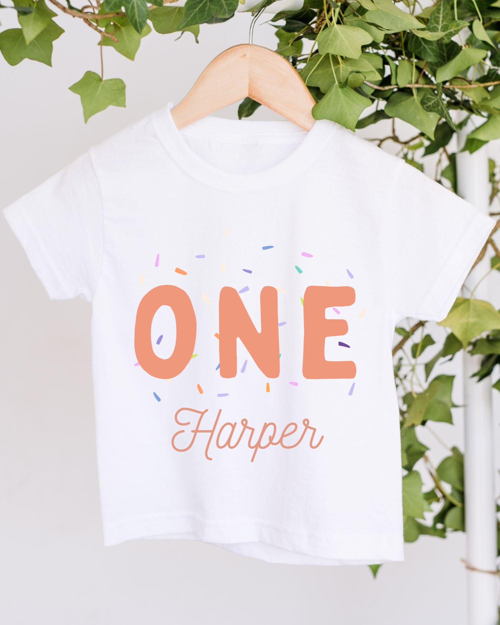 A white t-shirt with the text "ONE Harper" printed on it, along with colorful sprinkles. The t-shirt is hanging on a wooden hanger, and there are green leaves visible in the background.