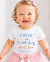 A smiling baby girl wearing a white t-shirt with text and a pink bow, standing in front of a white background.