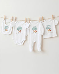 A row of white baby clothes with the text "Little Wicket Keeper" printed on them, along with a small baby bib.