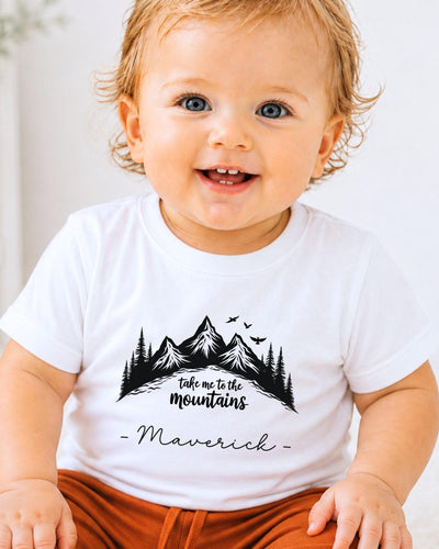 A smiling baby wearing a white t-shirt with a mountain graphic and the text "Take me to the mountains" and "Maverick" printed on it.