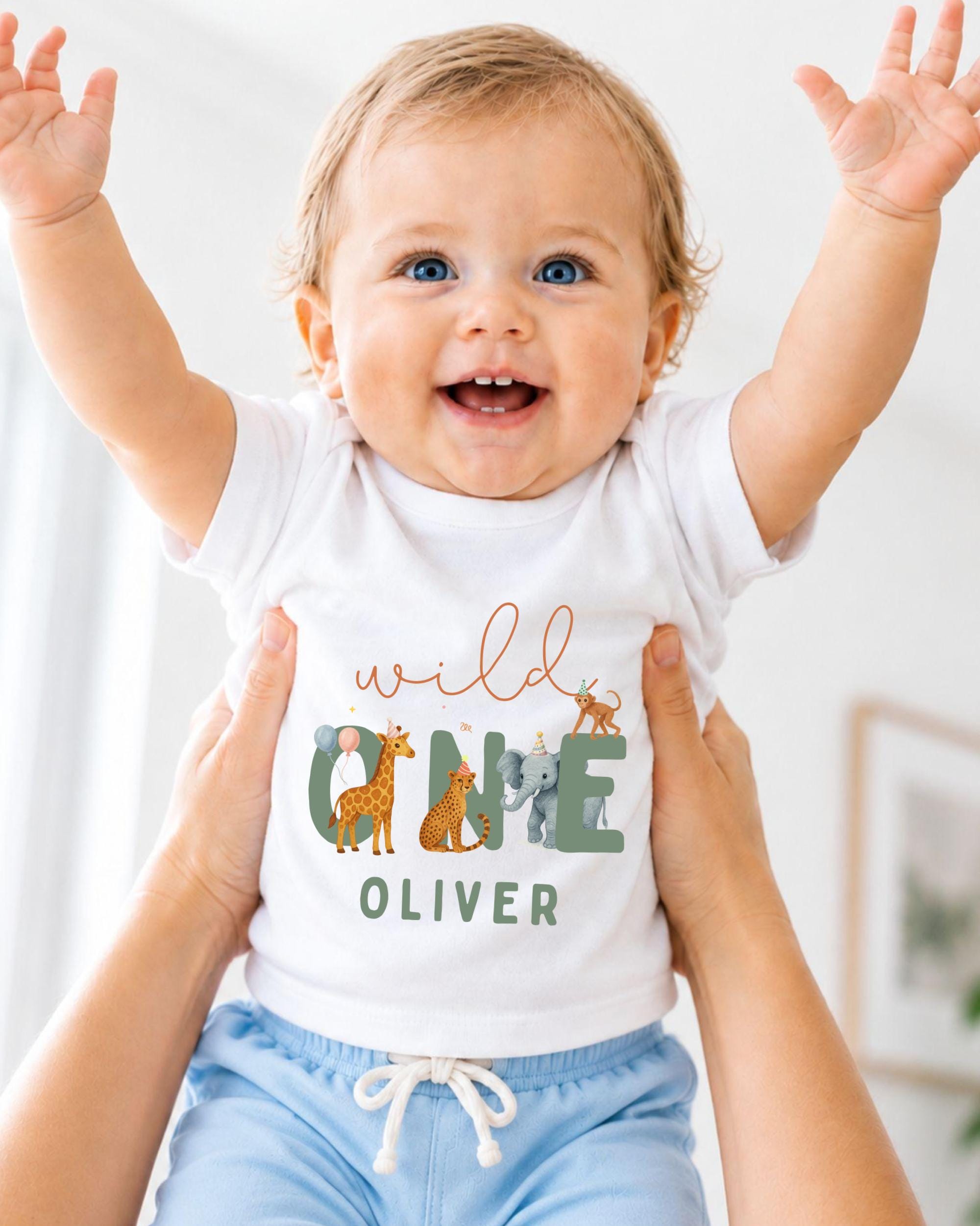A smiling baby wearing a white t-shirt with a graphic of animals and the name "Olivier" printed on it, being held up by someone's arms.