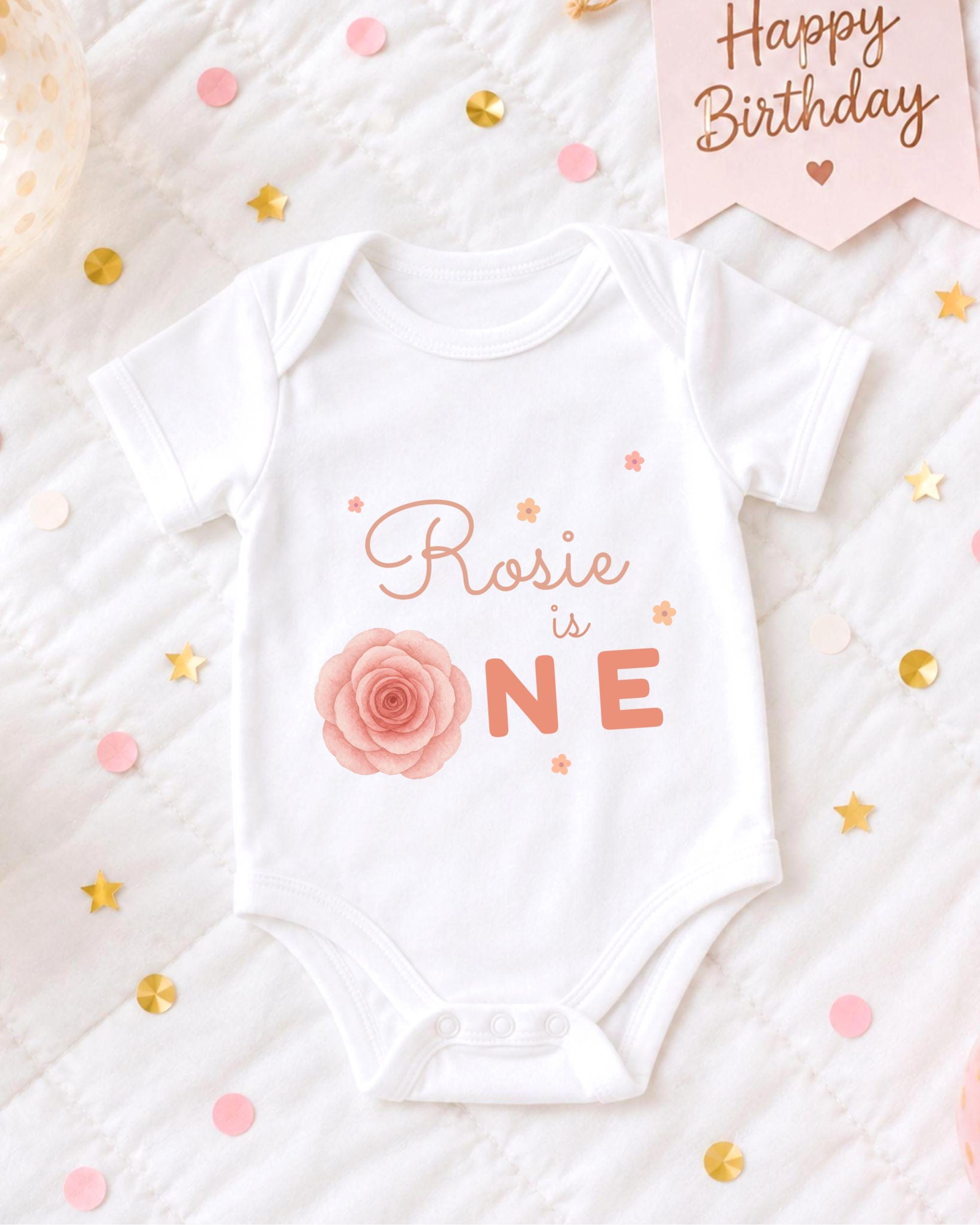 A white onesie with a rose design and the text "Rosa is one" printed on it, surrounded by confetti and a birthday card.