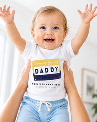 A smiling baby wearing a white shirt with a graphic and text, being held up by someone's arms.
