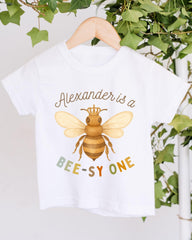 A white t-shirt with a printed design of a bee wearing a crown, along with the text "Alexander is a BEE-SY ONE" printed on it.