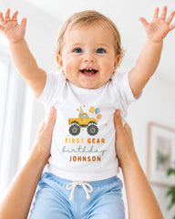 A smiling baby wearing a white t-shirt with a first birthday truck graphic and the text "FIRST GEAR birthday JOHNSON" is being held up by two people.