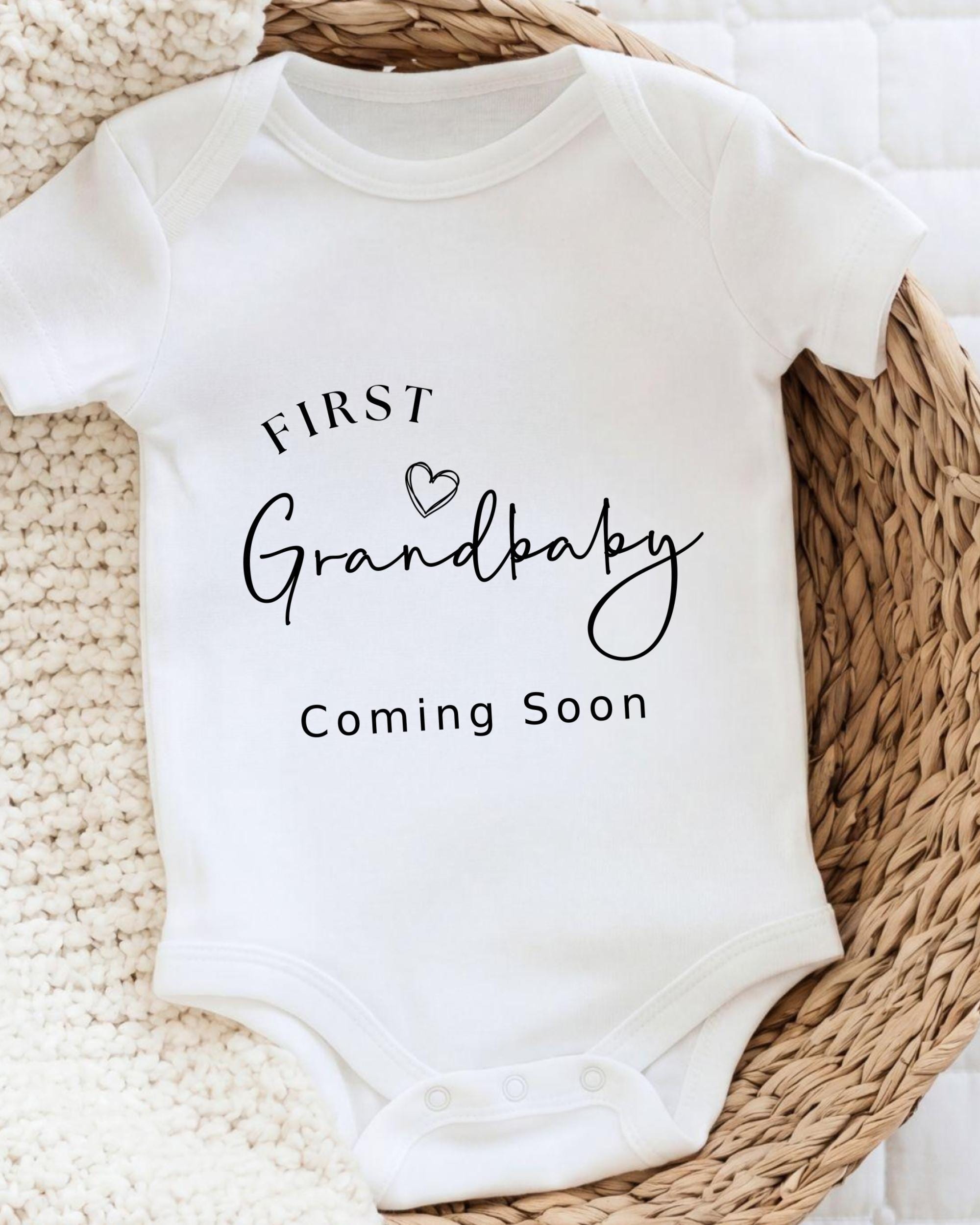 A white baby onesie with the text "FIRST Grandbaby Coming Soon" printed on it, placed on a woven basket.