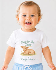A smiling baby boy wearing a white t-shirt with a cartoon image of a cow and the text "New to the Crew Peyton" printed on it.