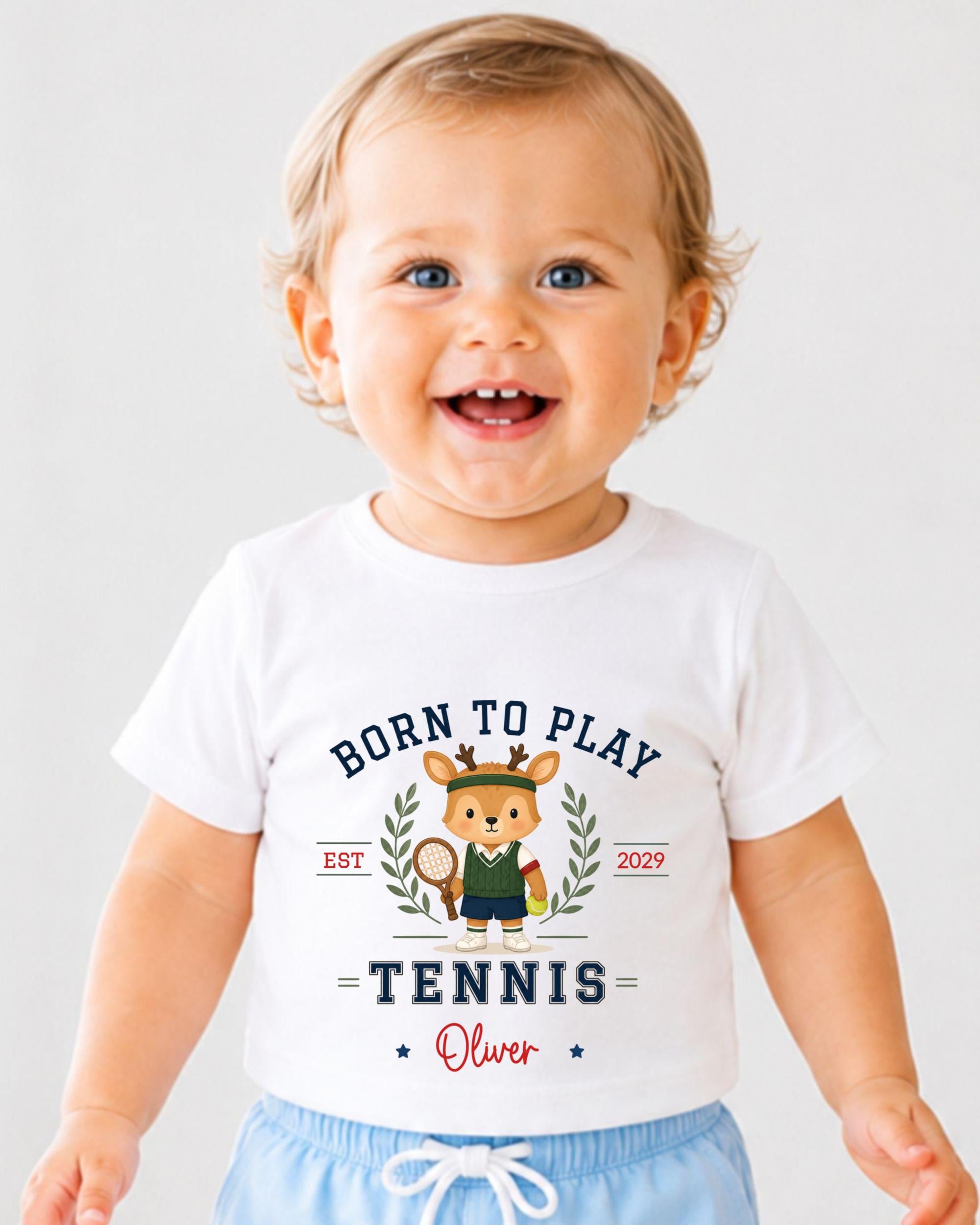 A smiling baby boy wearing a white t-shirt with a tennis-themed design and text.