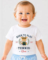 A smiling baby boy wearing a white t-shirt with a tennis-themed design and text.