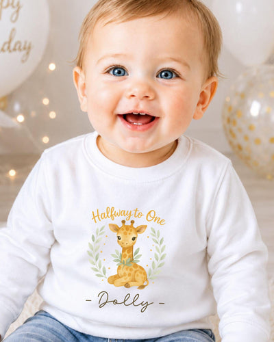 A smiling baby boy wearing a white shirt with a giraffe graphic and the text "Halfway to One - Dolly" printed on it.