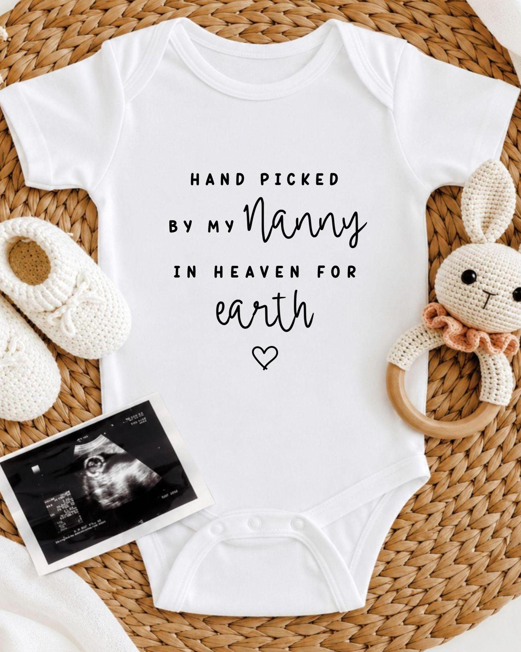 A white baby onesie with the text "HAND PICKED BY MY NANNY IN HEAVEN FOR EARTH" printed on it, along with a crocheted bunny toy and a black and white photograph of an infant's head.