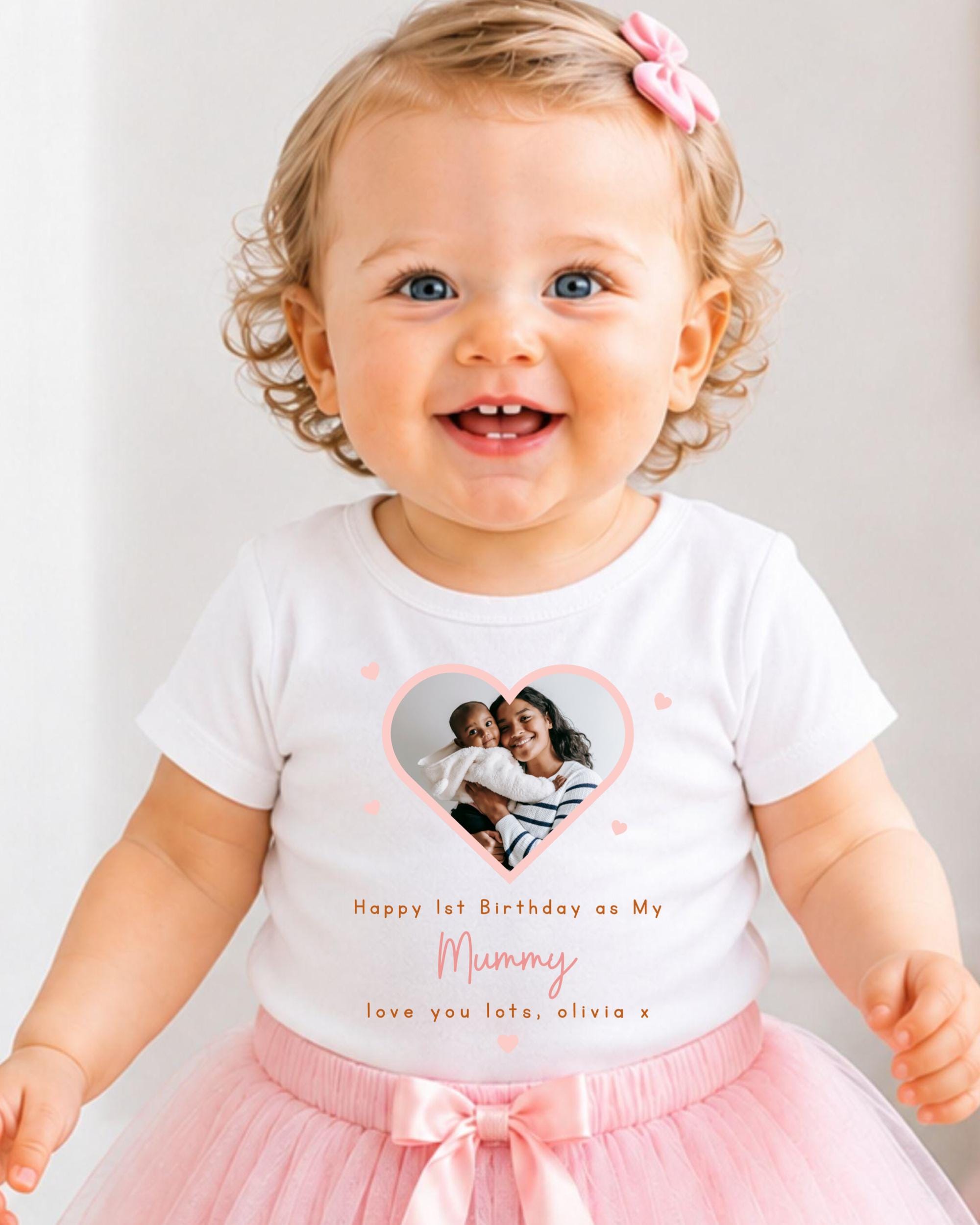 A smiling baby girl wearing a white t-shirt with a heart and a photo of her mother, and a pink tutu skirt.