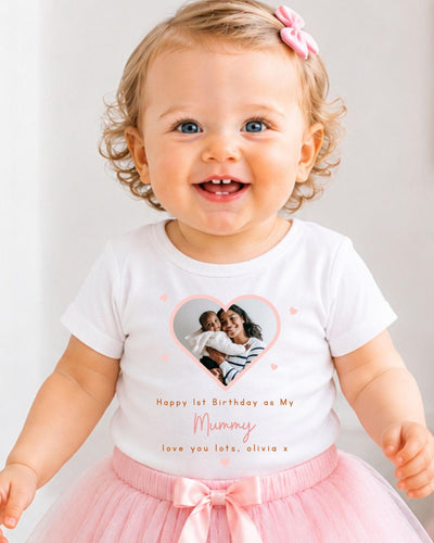 A smiling baby girl wearing a white t-shirt with a heart and a photo of her mother, and a pink tutu skirt.