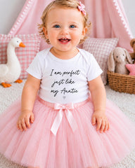 A smiling baby girl wearing a white t-shirt with text and a pink tutu, sitting on a pink carpet with stuffed animals in the background.