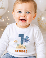 A smiling baby boy wearing a white shirt with a blue number 1 and an illustration of an octopus and a fish.