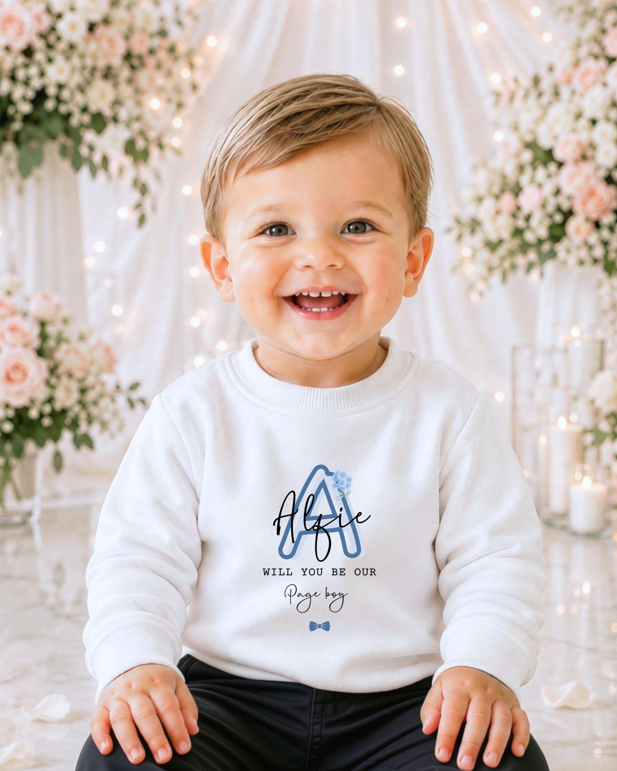 A smiling baby boy wearing a white sweater with a blue logo and text, sitting in front of a floral background with white lights.