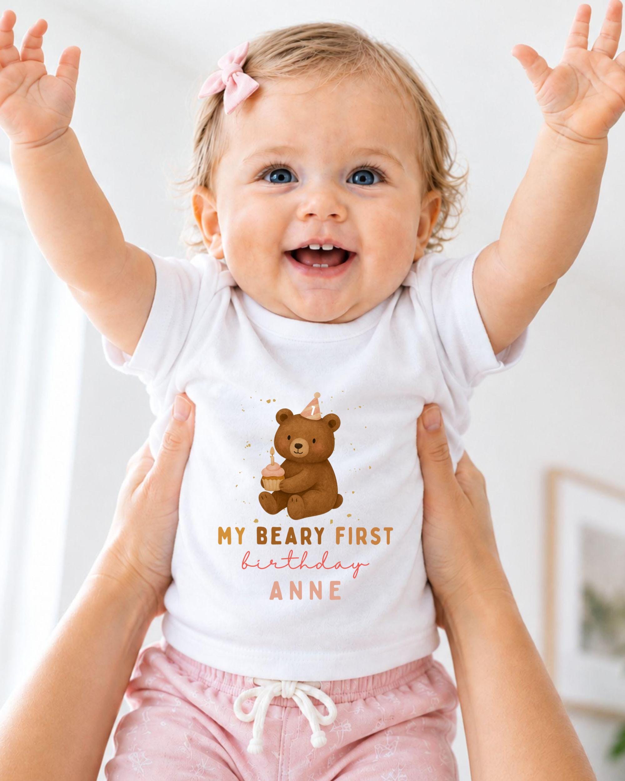 A smiling baby girl wearing a white t-shirt with a teddy bear and the text "My Beary First birthday Anne" is being held up by someone's arms.