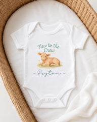 A white baby onesie with a printed image of a baby lamb and the text "New to the Crew Peyton" printed on it, placed on a white blanket.