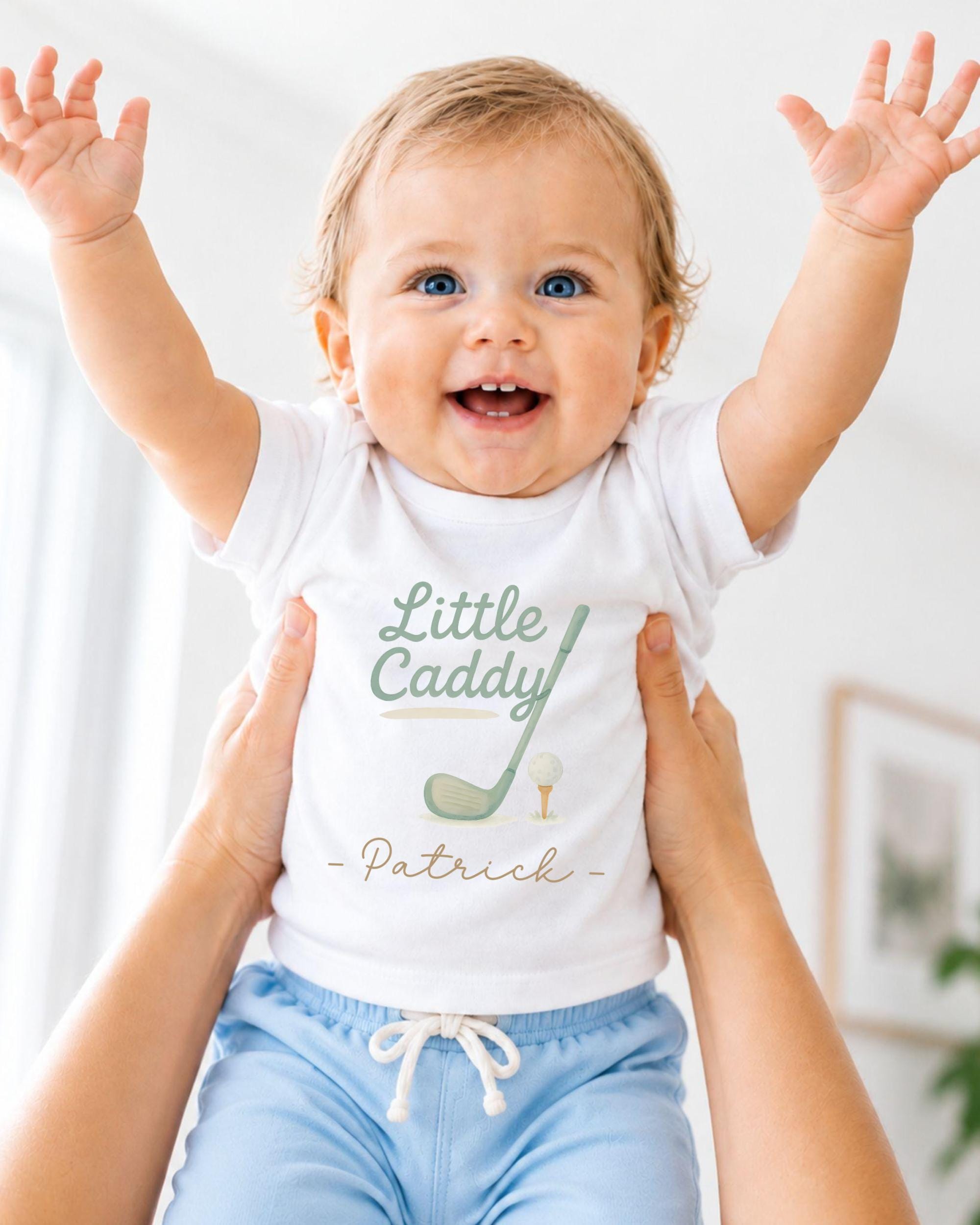 A smiling baby wearing a white t-shirt with a golf club and the name "Patrick" printed on it, being held up by someone's arms.