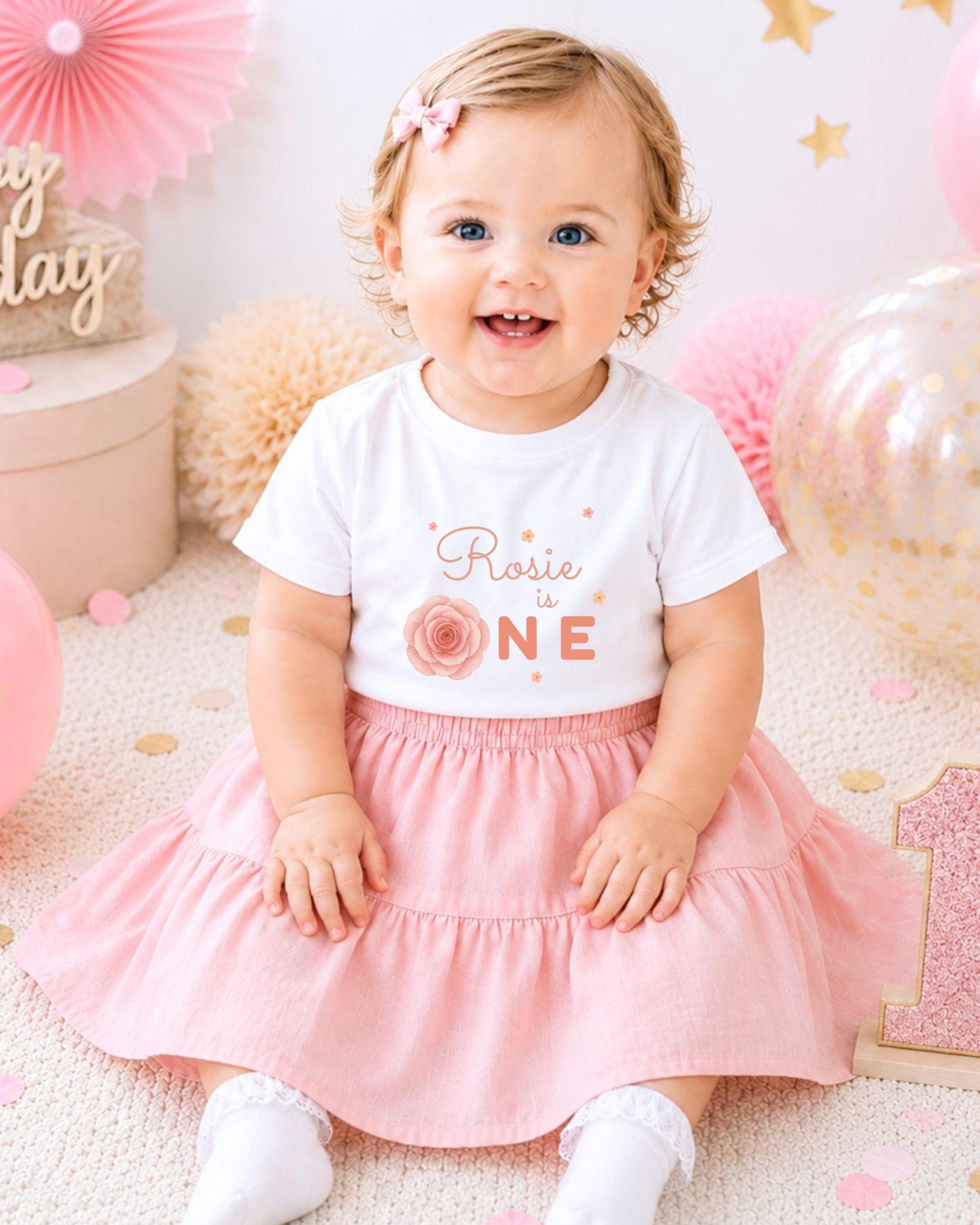 A smiling baby girl wearing a white t-shirt with a rose design and a pink skirt, sitting on a pink and white background with balloons and confetti.