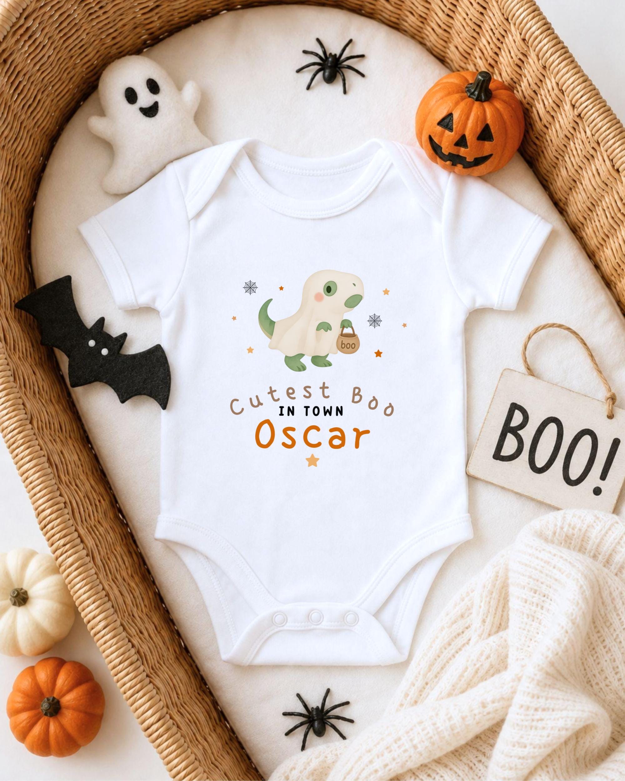 A white onesie with a cartoon dinosaur and the text "Cute Boo Oscar" is displayed on a bed, surrounded by Halloween decorations such as a ghost, a pumpkin, a bat, and a sign that says "BOO!".