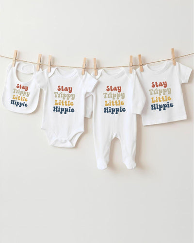 A row of four white baby clothes with the text "Stay Trippy Little Hippie" printed on them, hanging on a clothesline.