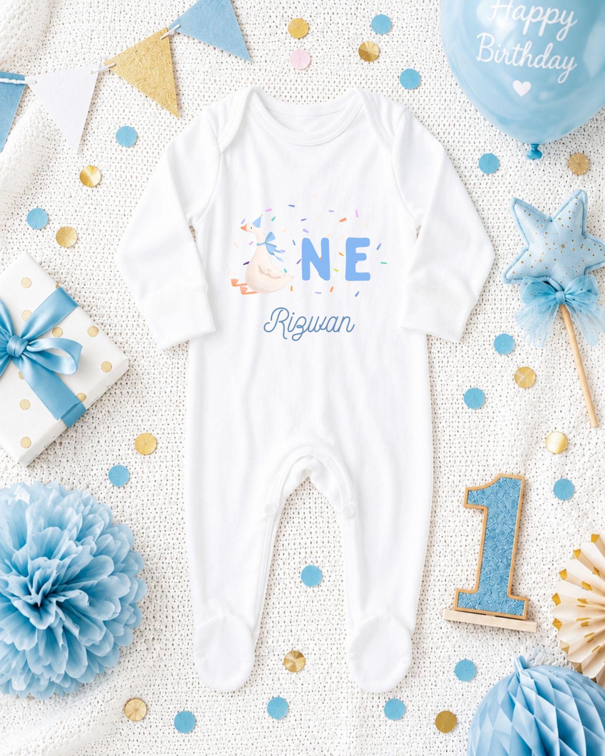 A white onesie with the name "Rigwan" printed on it, surrounded by birthday decorations such as a blue balloon, a blue and gold star, and blue and gold confetti.