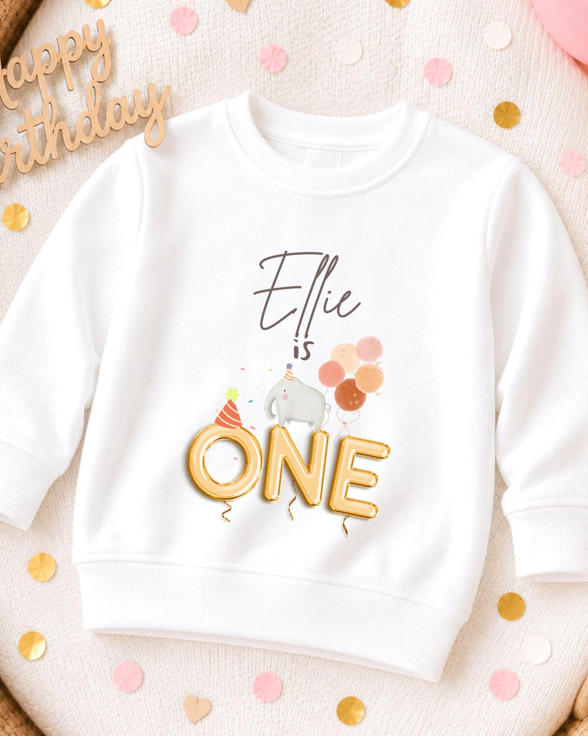 A white sweatshirt with a birthday-themed design, including a gold "One" and a small elephant wearing a party hat.
