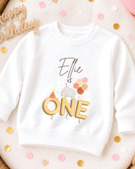 A white sweatshirt with a birthday-themed design, including a gold "One" and a small elephant wearing a party hat.