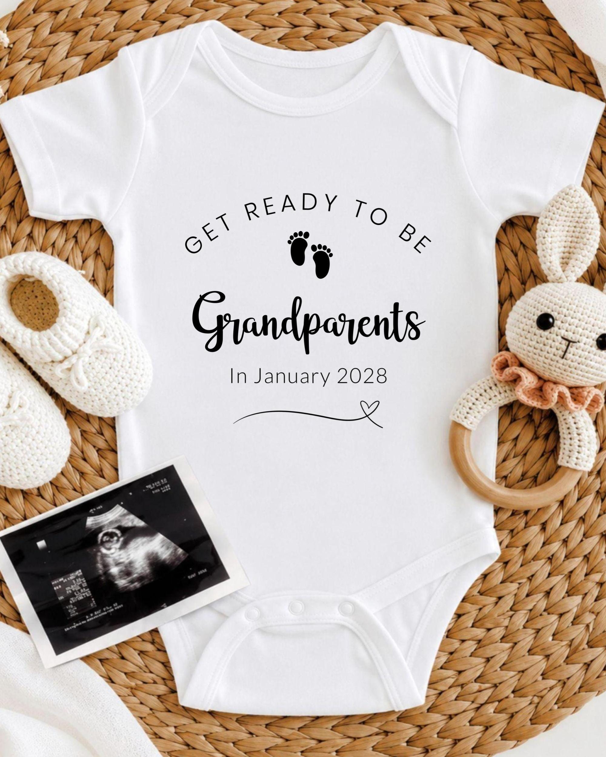 A white baby onesie with the text "GET READY TO BE Grandparents In January 2028" printed on it, along with a pair of white crocheted shoes and a stuffed rabbit toy. There is also an Ultrasound image of a baby's feet visible in the image.