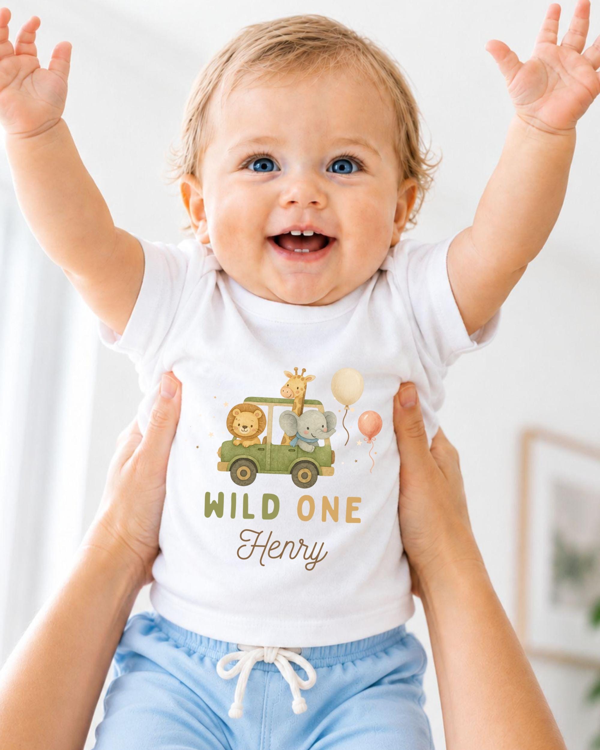 A smiling baby wearing a white t-shirt with a cartoon car and animals, being held up by someone's arms.