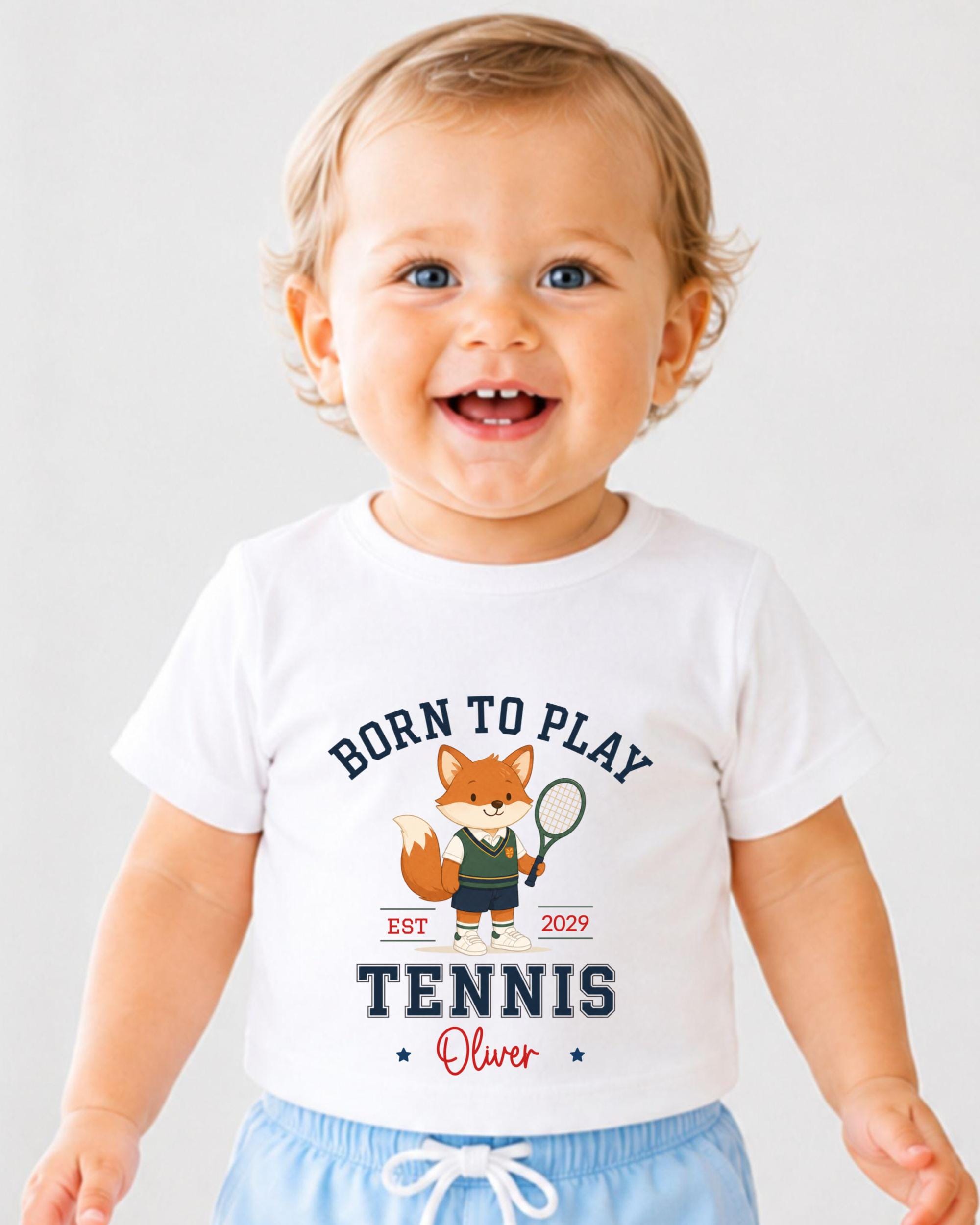 A smiling baby boy wearing a white t-shirt with a tennis-themed graphic and text.